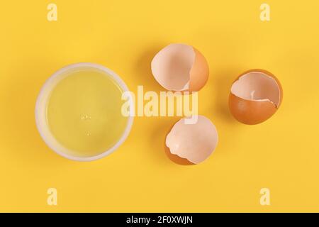 Bowl with egg white next to broken eggshells on yellow background Stock Photo