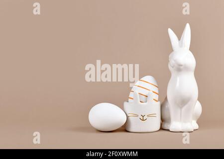 Golden striped Easter egg in bunny egg cup next to rabbit figure and white egg on side of beige background with empty copy space Stock Photo