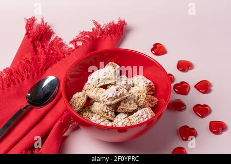 romantic or valentine breakfast with red bowl of frosted wheat cereal, napkin and spoon with red hearts table scatter on a pale pink background Stock Photo