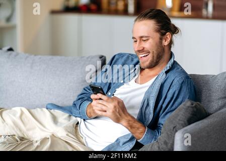 Satisfied attractive guy is sitting at the couch, using smartphone, spend time in the network. Smiling guy got a good news or message, browsing internet, chatting online with friends or family Stock Photo