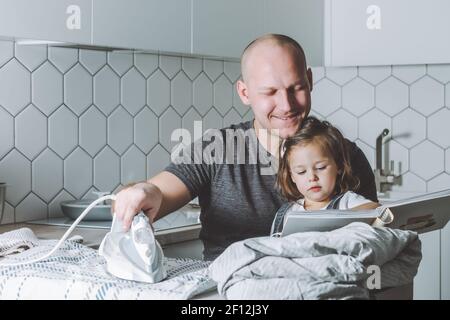 Man ironing bed linen and reading book to his little daughter. Father is engaged in household chores. Stock Photo