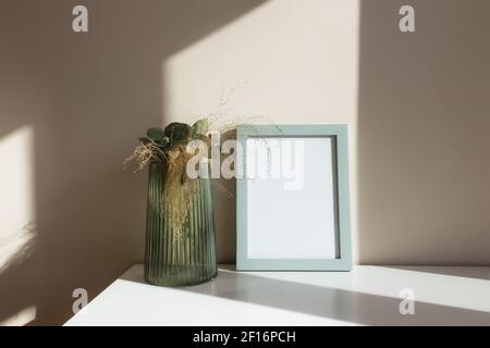 Glass vase with herbarium flowers, eucalyptus branches, empty white photo frames on the white table in the interior with beige walls near window. Stock Photo