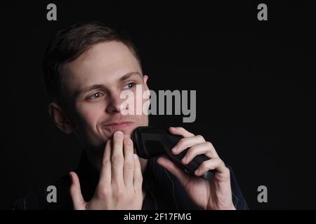 A young man shaves his face with an electric razor. Stock Photo