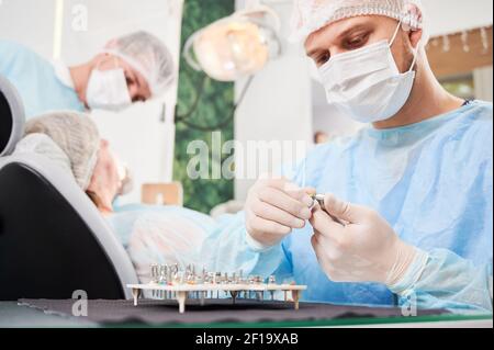 Dentist on foreground, wearing disposable sterile clothes, putting surgical nozzle on drill before implant installation. Patient and assistant on blurred background. Concept of dentistry. Stock Photo