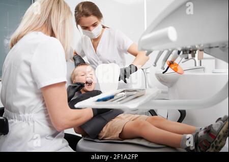 Little boy sitting in dental chair while two female dentists checking kid teeth. Dentist examining boy teeth with dental instrument. Concept of pediatric dentistry and dental care. Stock Photo