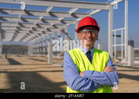Happy Manager in Red Hardhat, Blue Shirt and Yellow Vest Looking at Camera. Portrait of Construction Foremen on Building Site. Stock Photo