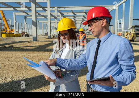 Businessperson Pointing at Blueprint. Man and Woman In Protective Clothing Discuss a Construction Project on Site. Stock Photo