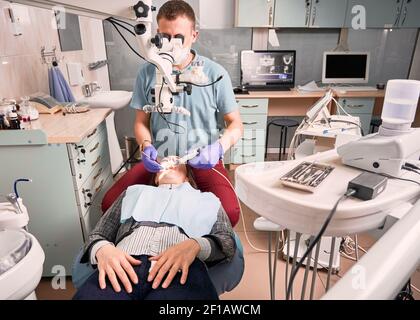 Young male dentist using microscope and dental instruments while treating woman teeth. Female patient lying in dental chair at modern medical center. Concept of dentistry, stomatology and dental care. Stock Photo