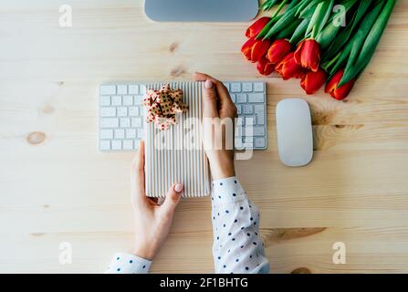 Top view of female elegant hands holding gift wrapping box over computer keyboard at office workplace. Delivery of gifts and internet orders. Stock Photo