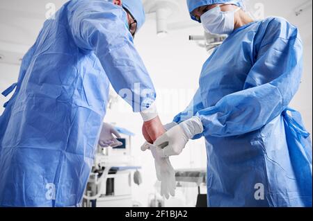 Assistant in blue surgical suit helping surgeon to put on sterile gloves. Two doctors in protective medical masks preparing for plastic surgery in clinic. Concept of medicine and surgery preparations. Stock Photo
