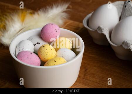 Eggs for Easter. Colorful chocolate Easter eggs in a bowl and raw eggs in egg carton. Feathers on wooden table. Stock Photo