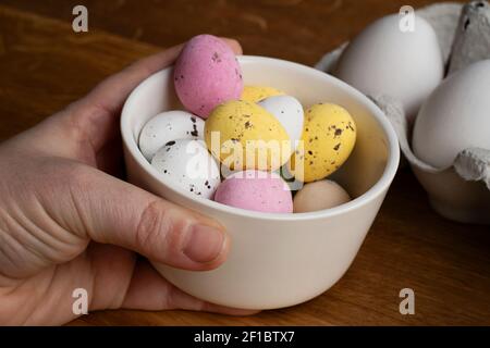 Hand holding up a bowl filled with chocolate Easter eggs in various colors. In the background is an egg carton on wooden table. Stock Photo