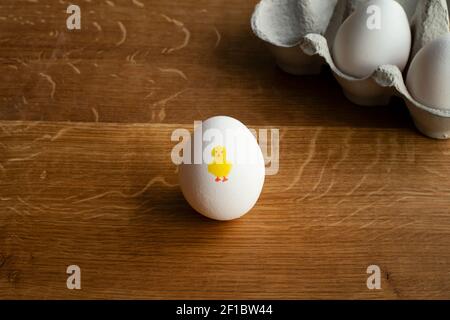 Cute Easter chicken painted on white egg on wooden table next to egg carton. Stock Photo