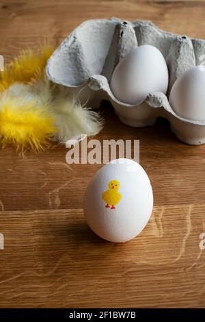 Fun Easter chicken painted on white egg placed in front of white and yellow feathers and egg carton on the breakfast table. Stock Photo