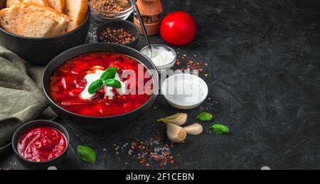 Fresh, delicious borscht in a deep black plate, on a background of bread, spices, garlic and sour cream on a black background. Panorama with space to Stock Photo