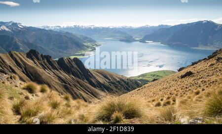 Beautiful vista with dry tussocks,lush green meadows,blue lake and snowy mountains wide shot made during sunny day on Isthmus Peak summit,New Zealand Stock Photo