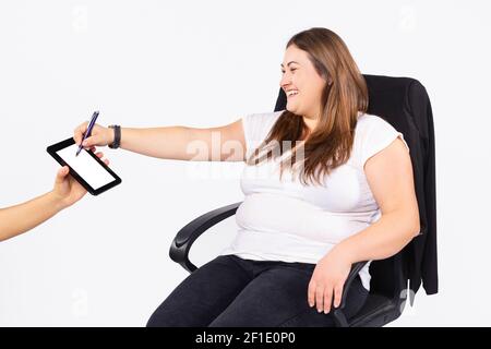 An overweight woman sits in a black office chair and paints on the bar that her hand gives her from her side on a white studio background. Stock Photo