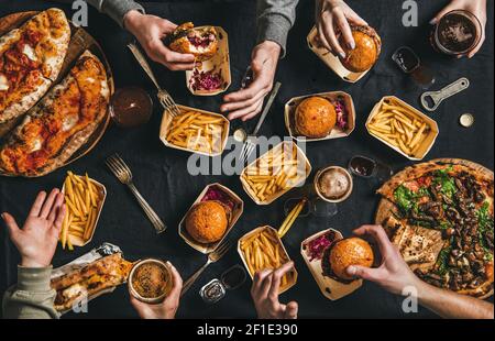 Lockdown fast food dinner from delivery service. Flat-lay of peoples hands eating burger, fries, sandwiche, pizza, drinking beer over table background, top view. Quarantine home party, takeaway food Stock Photo