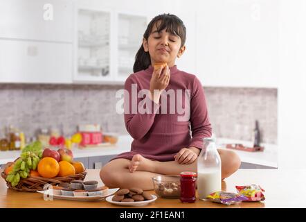 A YOUNG GIRL SITTING IN KITCHEN AND PLAYFULLY TASTING A PIECE OF CAKE Stock Photo