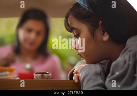 A YOUNG GIRL SITTING AND STARING AT A CUPCAKE WITH MOTHER SITTING IN THE BACKGROUND Stock Photo