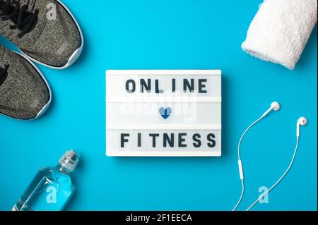 Online fitness - text on display lightbox on blue background. Flat lay - Sneakers, a bottle of water, white headphones. Sport concept. Stock Photo