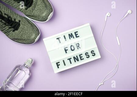 Time for fitness - motivanional text on display lightbox on purple background. Flat lay - Sneakers, a bottle of water, white headphones. Sport concept Stock Photo
