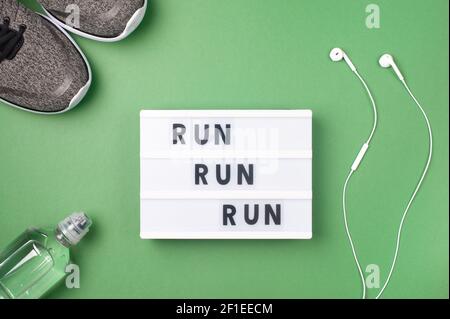 Run Run Run - motivanional text on display lightbox on green background. Flat lay - Sneakers, a bottle of water, white headphones. Sport concept. Stock Photo