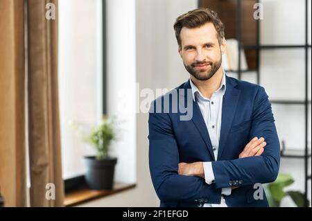 Successful confident young bearded brunet adult male entrepreneur businessman professional in business casual clothes standing with arms crossed folded at the modern home office and looking at camera Stock Photo