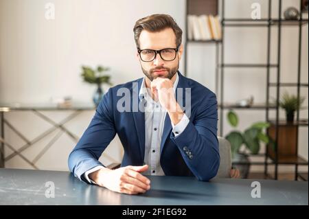 Portrait of sophisticated young bearded confident entrepreneur man in business casual clothes sitting at the desk and looking at camera, placed hand on his chin and thinking. Copy space concept Stock Photo