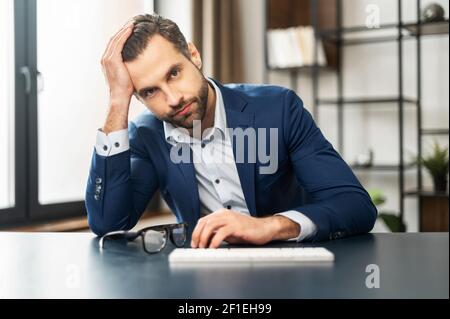 Helpless unlucky young handsome businessman with beard sitting at the desk with one hand on keyboard, another supporting head, feeling upset from unexpected problem bad news, lost money or business Stock Photo