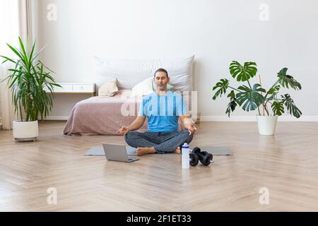 Meditation practice - middle aged man during online yoga steaming at home. A man sits on a yoga mat in front of a laptop monitor and meditates. Stock Photo