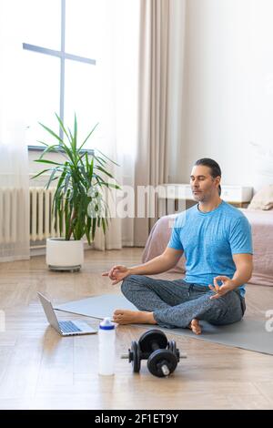 Vertical photo. Meditation practice - middle aged man during online yoga steaming. A man sits on a yoga mat in front of a laptop monitor and meditates Stock Photo