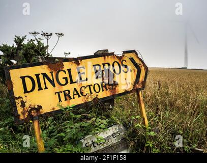 rusting sign beside corn field ,Dingle brothers tractors pointing to  wind turbine in the mist Stock Photo