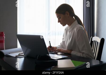Focused young woman sitting at table with opened laptop and taking notes while working at home. Modern technology and people concept. Stock Photo