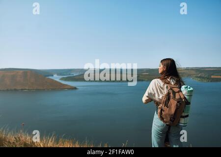 Happy young woman with backpack standing on high hill and looking at wide Dniester river. Pretty lady walking on fresh air at national park Podillya Tovtry Stock Photo