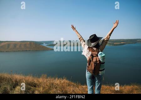 Young female tourist in cowboy hat holding hands up while standing on mountain peak near Bakota bay. Concept of travelling and adventure. Stock Photo