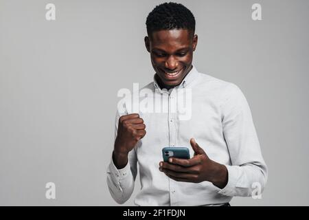 Handsome excited young african business man using mobile phone isolated over gray background, wearing white formal shirt, celebrating success Stock Photo