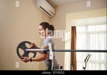 A young fit woman puts a metal disc on barbell during bodybuilding workout. Exercising at home. Stock Photo