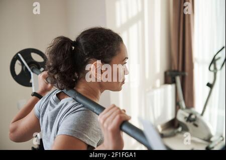 Rear view of African american young sporty woman exercising heavy duty squat with barbell in home gym. Heavy weight training. Stock Photo