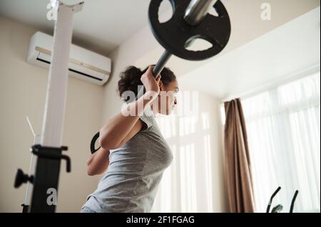 Back view to African young sporty woman doing heavy duty squat in home gym with barbell. Bodybuilding. Stock Photo