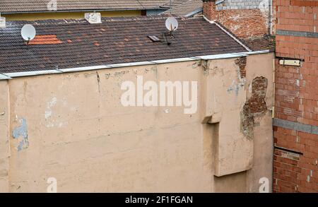 Old residential building wall, paint peeling, some bricks visible under plaster, tv satellite dishes on roof Stock Photo