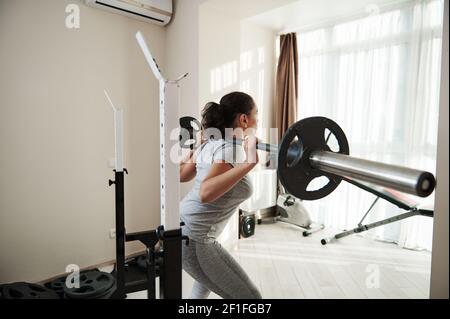 African American young sporty woman doing heavy duty squat in home gym with c barbell. Heavy weight training Stock Photo