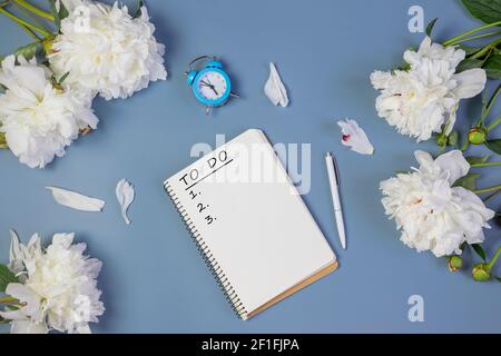 open notebook for writing plans, to-do list or dreams with fresh peonies, alarm clock, on light blue background Stock Photo