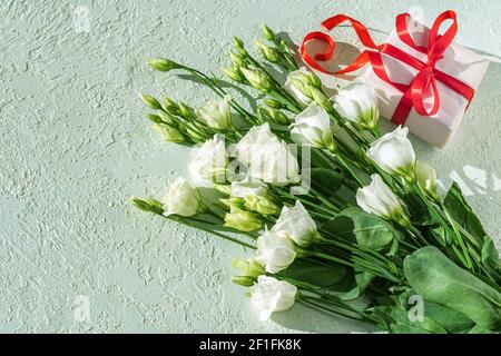 White delicate small roses and a white gift with a red ribbon on a light plaster background, copy space, top view, banner. Greeting card for mother's Stock Photo