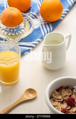 Aerial view of cereal bowl, spoon, oranges, juicer, cloth, jug with milk and glass with orange juice, on white wood, vertical Stock Photo