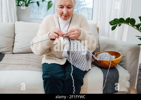 An elderly woman of seventy years old is embroidering with knitting needles while sitting on the sofa. Stock Photo