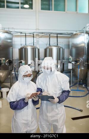 Vertical portrait of two young workers wearing hazmat suits while using digital tablet at modern chemical factory Stock Photo