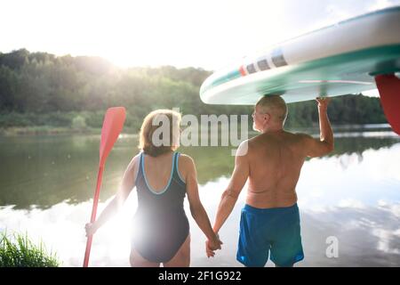 Rear view of senior couple carrying paddleboard by lake in summer. Stock Photo