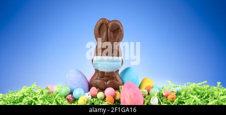 Chocolate Easter bunny wearing a medical mask surrounded by painted Easter eggs and colorful decorations. Social distancing Easter celebrations due to Stock Photo