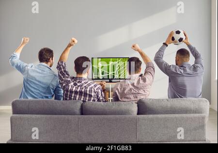 Four adult male friends with arms raised emotionally watching a football match on TV at home. Stock Photo